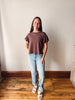 A woman stands in a room with a beige wall and wooden floor, wearing a short-sleeve ruffled-sleeve top in a muted brownish-purple color, light-wash blue jeans, and white slide sandals.
