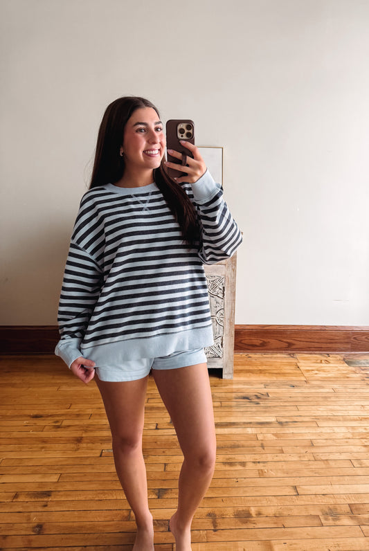 A woman takes a mirror selfie wearing a blue and white striped long-sleeve crewneck sweatshirt and light blue shorts in a room with a wooden floor and off-white wall.