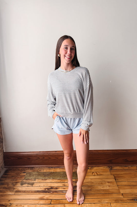 A woman stands barefoot in a room with a light beige wall and wooden floor, wearing a long-sleeve blue and white striped crewneck sweatshirt and light blue shorts.