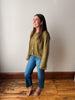 A woman wearing a textured olive green knit cardigan with gold buttons, blue jeans, and flat sandals, standing on a wooden floor against a plain off-white wall.