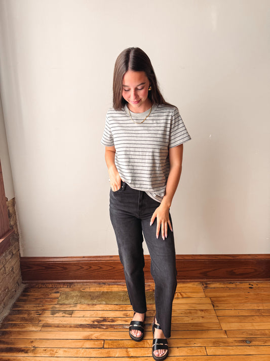 A woman modeling a short-sleeve crewneck t-shirt with thin gray horizontal stripes, paired with dark gray slim-fit jeans and black slide sandals, standing on a wooden floor against a plain beige wall.