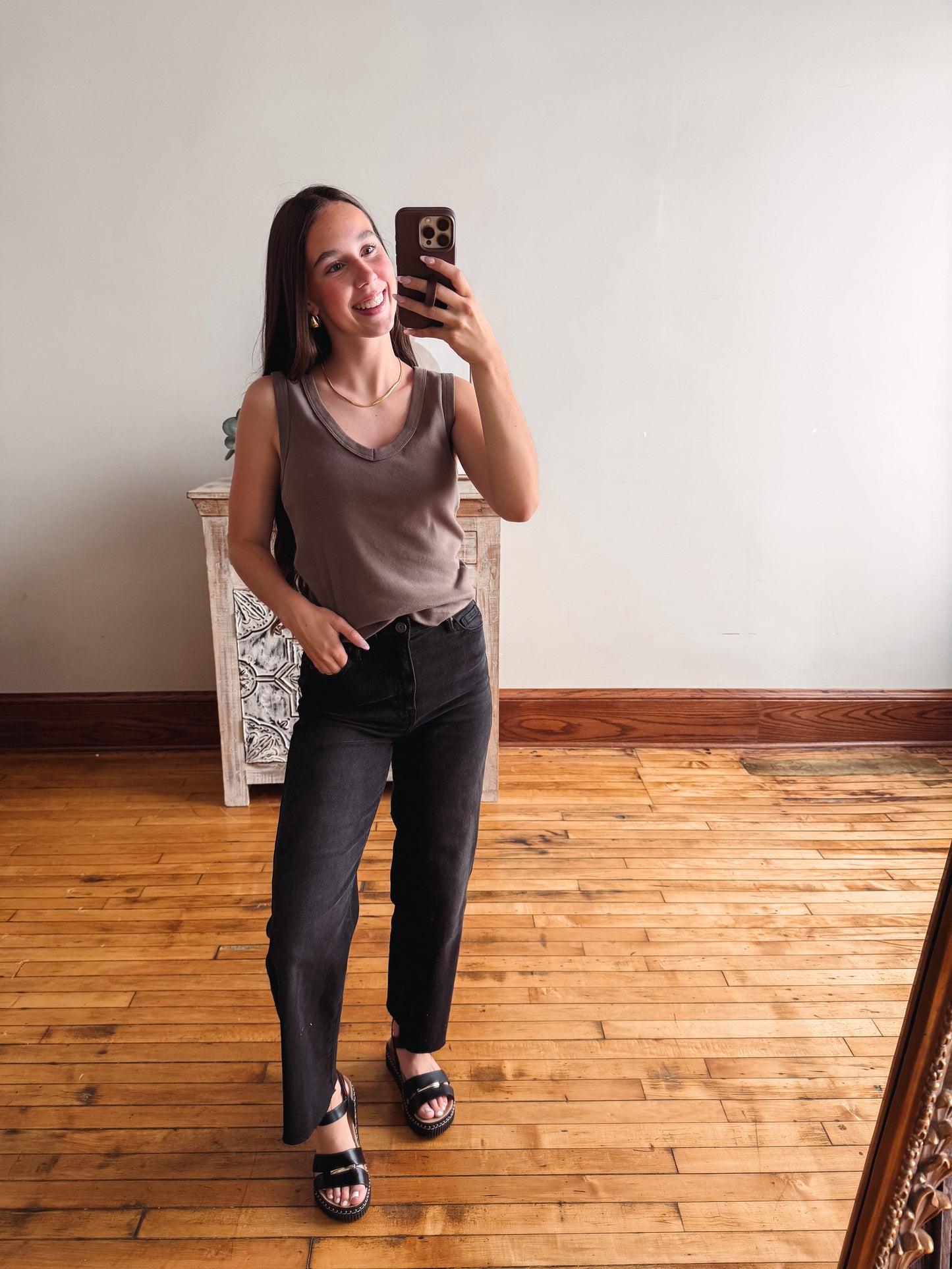 A woman takes a mirror selfie in a room with hardwood floors, wearing a brown sleeveless top, dark charcoal gray straight-leg jeans, and black sandals.