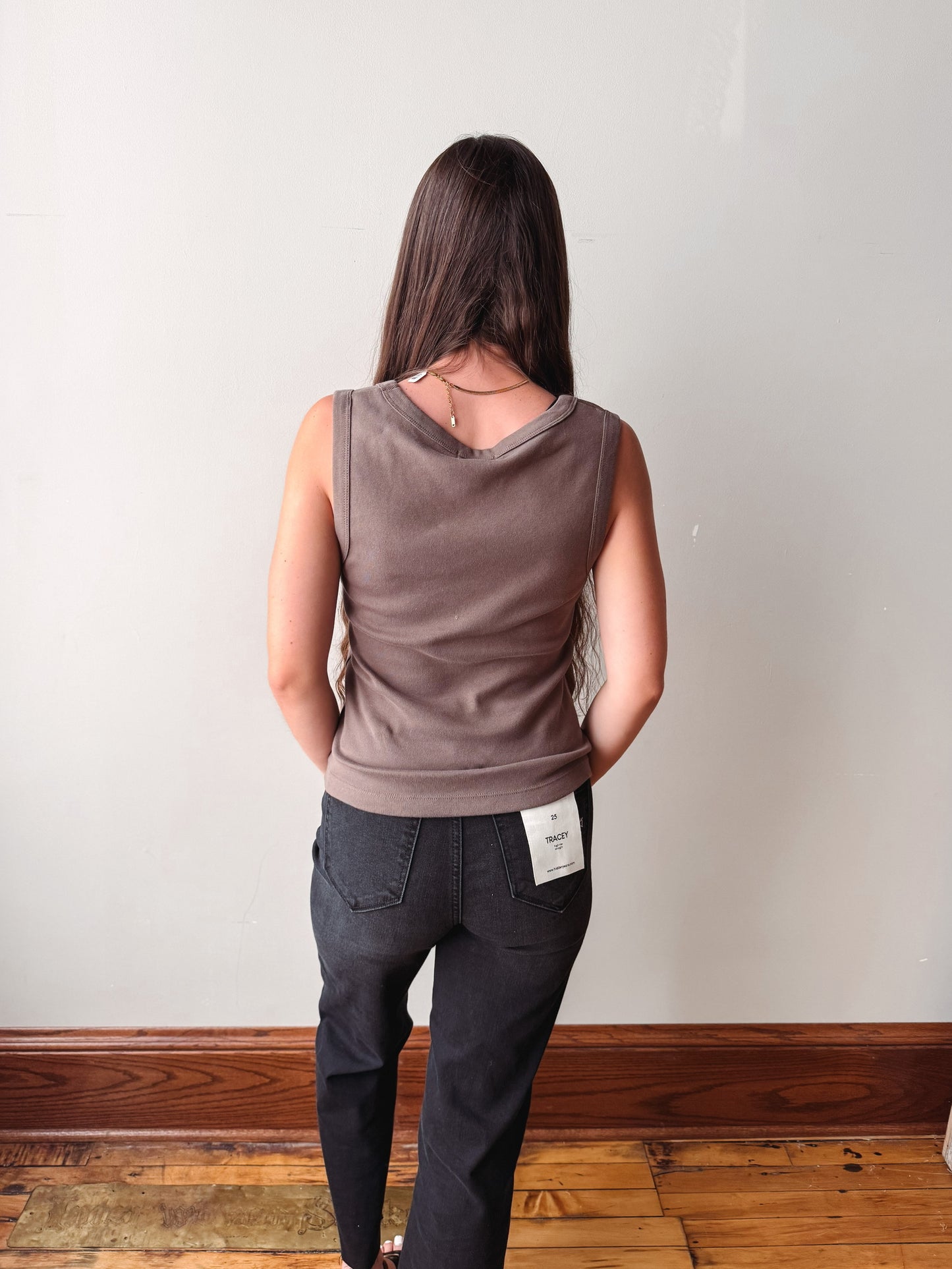 A woman viewed from behind wearing a sleeveless, cowl-neck taupe top and dark gray jeans, standing on a wooden floor against a plain off-white wall.