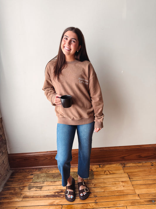 A woman stands indoors against a plain off-white wall on a wood floor, wearing a tan crewneck sweatshirt with a small embroidered logo on the left chest, blue jeans, and black slide sandals while holding a black mug.