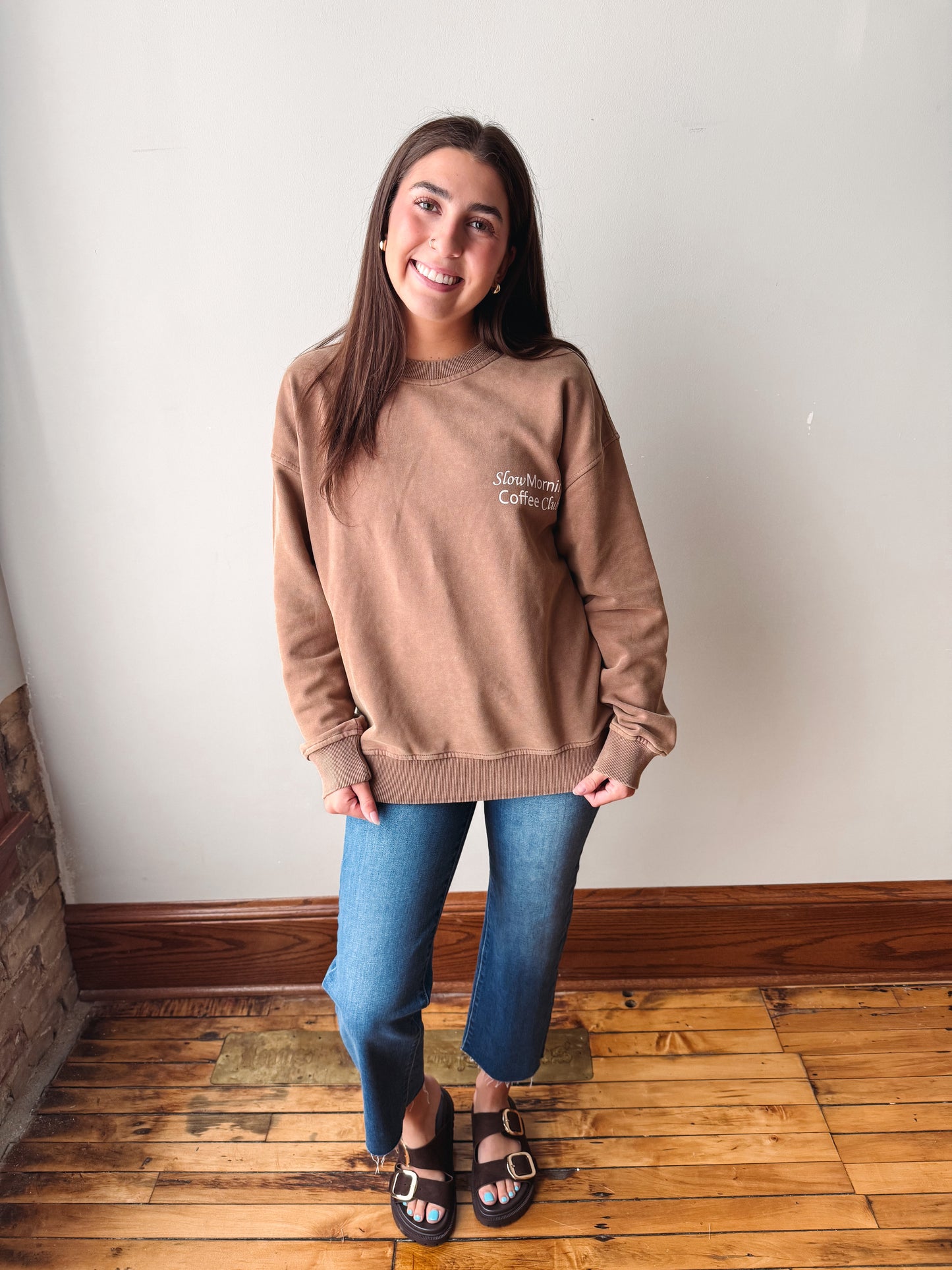 A woman stands indoors wearing a taupe crewneck sweatshirt with 'Slow morning coffee club' printed on the chest, blue jeans, and brown sandals, posing against a white wall and wooden floor.
