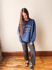 A woman wearing a blue crewneck sweatshirt with white text that reads 'NO PLANS Sunday Club MEMBERS ONLY', paired with dark gray wide-leg pants and black sandals, standing on a wooden floor against a beige wall.