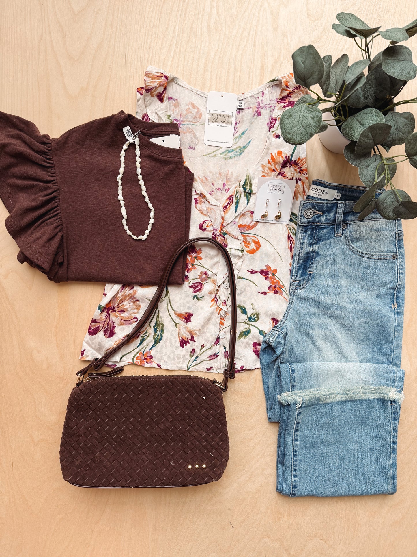 A styled flat-lay outfit on a light wood background featuring a dark brown ruffled top, a cream floral blouse, light blue rolled-calf jeans, a dark brown woven crossbody bag, a white beaded necklace, and a potted eucalyptus plant.