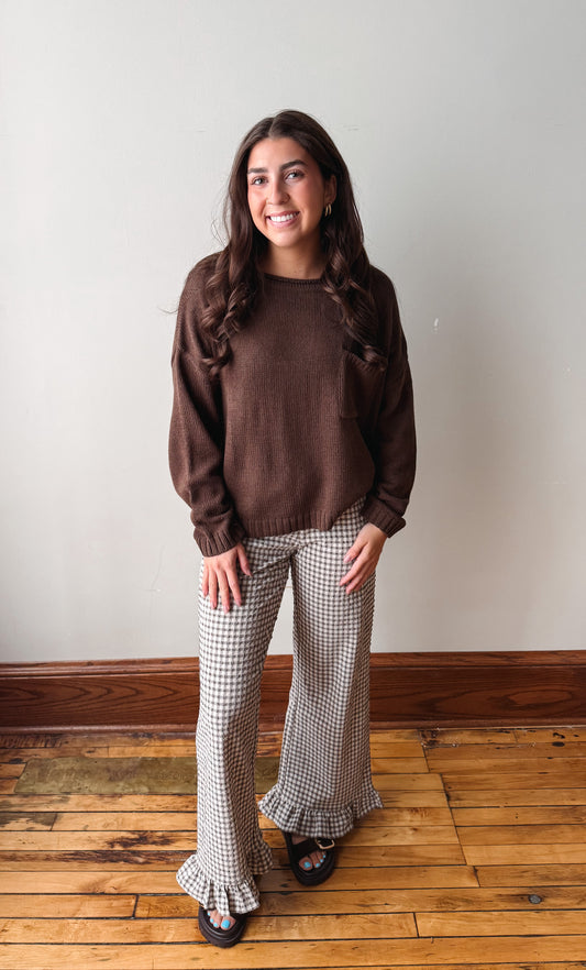 A woman stands in a room with a light gray wall and wooden floor, wearing a brown knit crewneck sweater and beige-and-black gingham flared pants with ruffled hems.