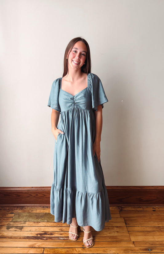 A woman modeling a dusty blue maxi dress with short flutter sleeves, a gathered sweetheart neckline, and side pockets, standing on a wooden floor against a plain beige wall.