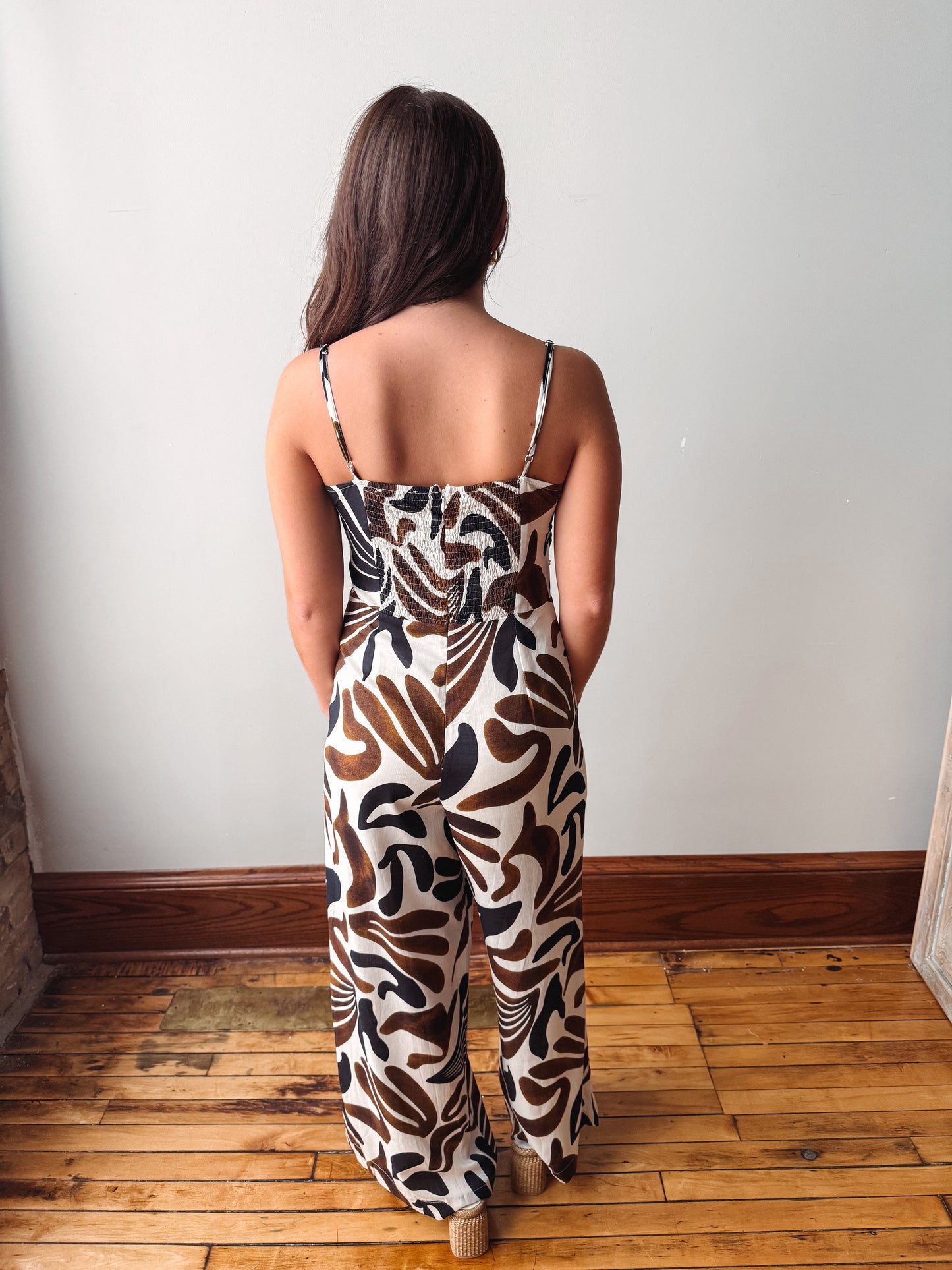 A woman viewed from behind wearing a sleeveless wide-leg jumpsuit with a brown, black, and beige botanical leaf pattern, standing on a wooden floor against a plain white wall.
