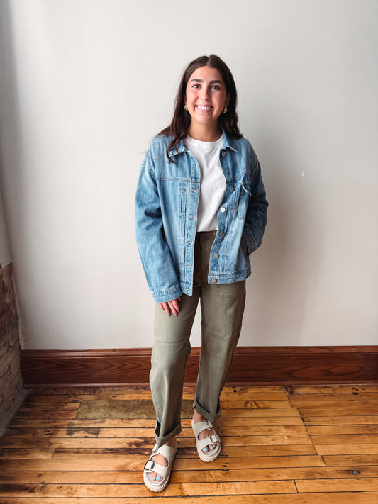 A woman stands indoors wearing a light blue denim jacket over a white top, olive green pants, and white slide sandals, photographed against a plain off-white wall with a wood floor.