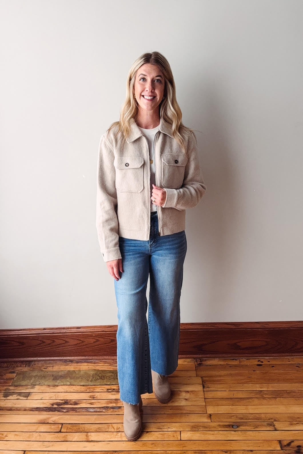 Woman wearing a beige jacket and blue jeans standing against a plain wall.