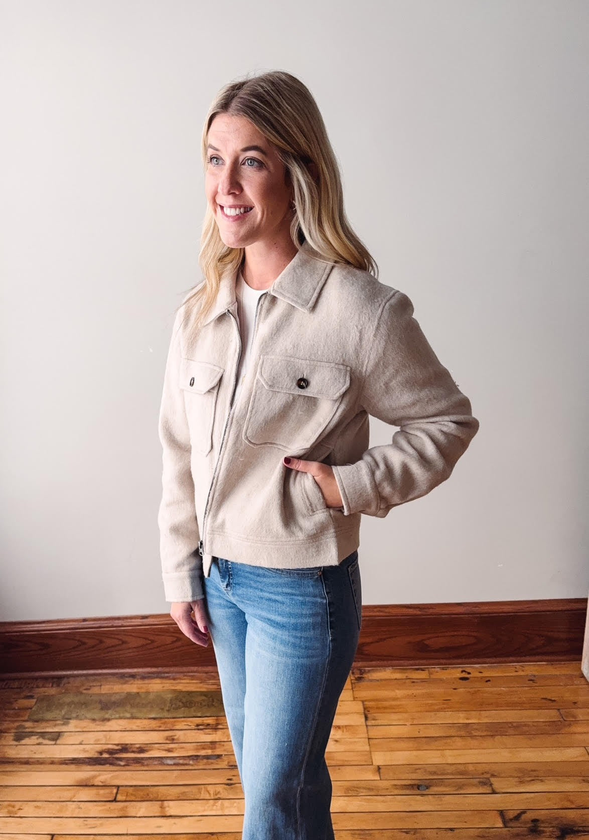 Woman wearing a beige jacket and blue jeans standing in a room with wooden flooring.