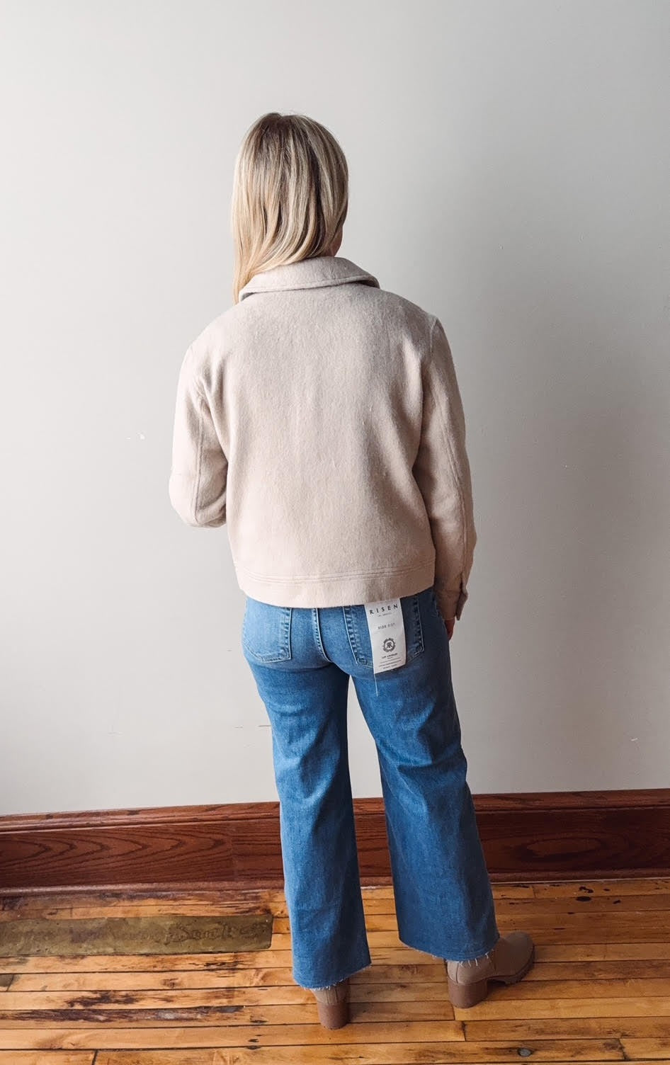 Person wearing a beige jacket and blue jeans standing against a plain wall.