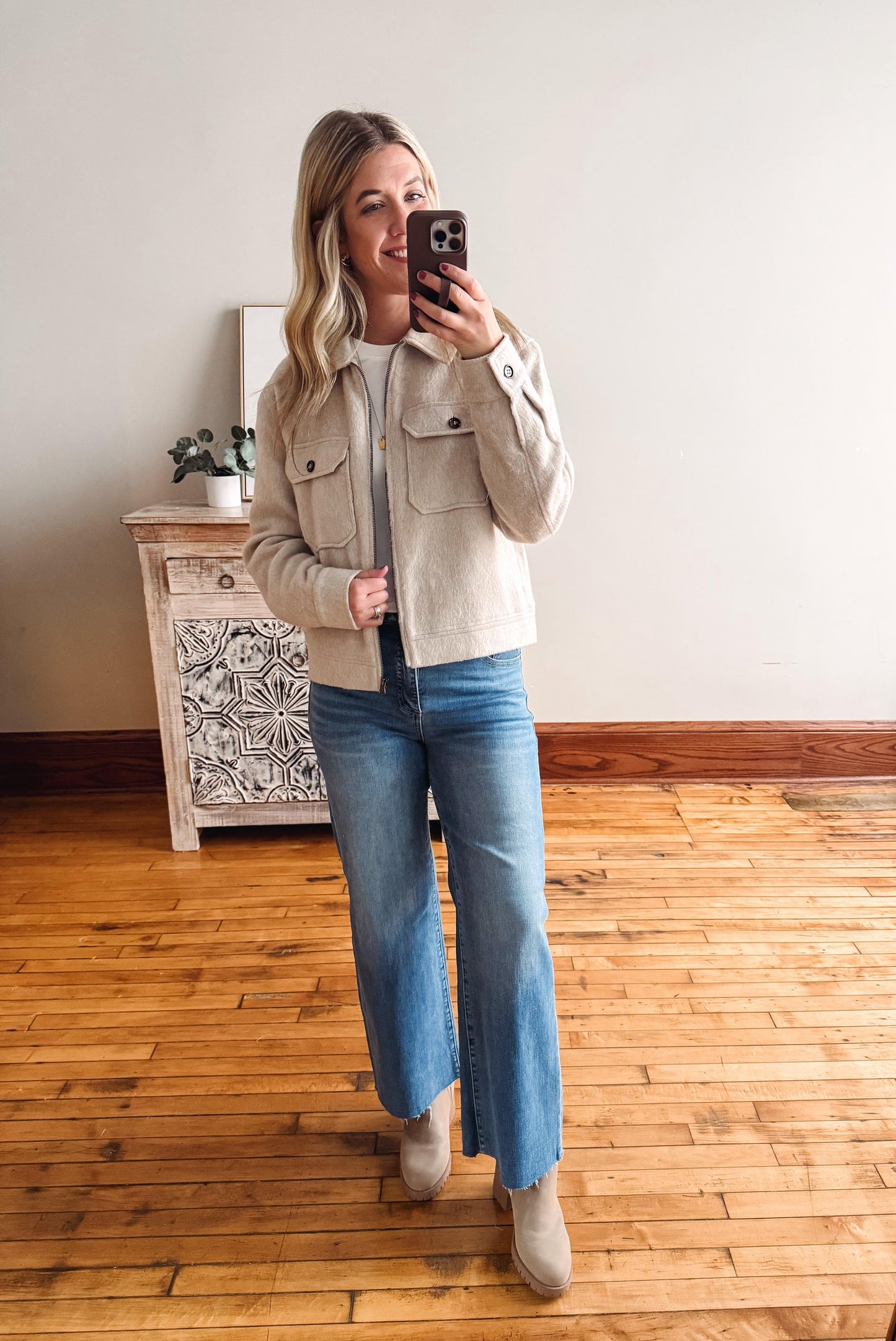 Woman taking a mirror selfie wearing a beige jacket and blue jeans in a room with wooden flooring.