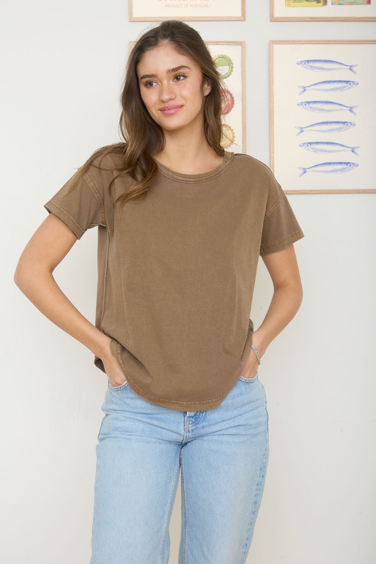 Woman wearing a brown t-shirt and light blue jeans standing against a white wall with framed artwork.