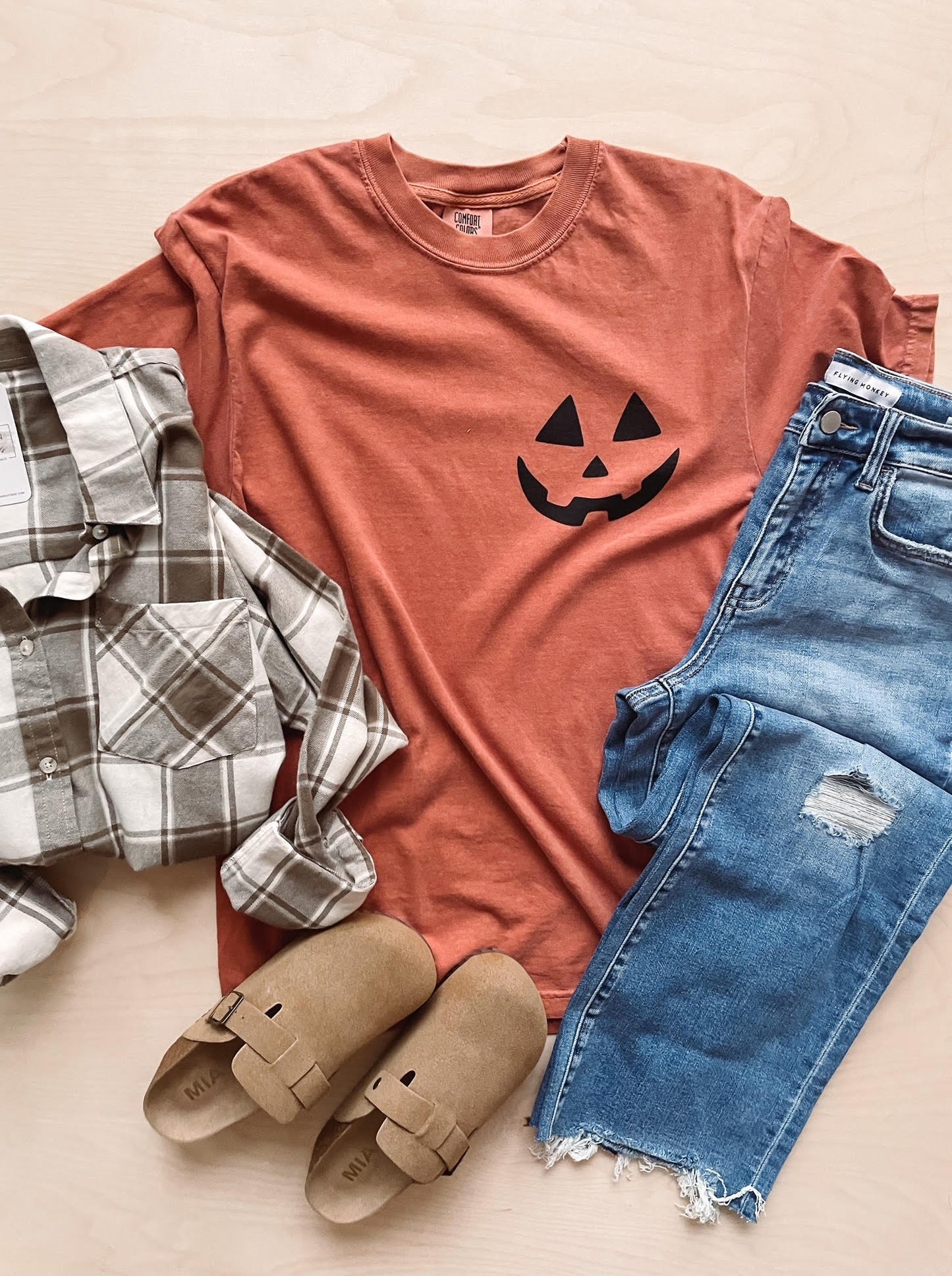 A yam colored short-sleeved graphic tee with a small black jack-o-lantern face on the right chest, displayed flat with a pair of blue denim jeans and a checkered patterned shirt on top, and a pair of tan sandals positioned in front.
