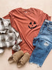 A yam colored short-sleeved graphic tee with a small black jack-o-lantern face on the right chest, displayed flat with a pair of blue denim jeans and a checkered patterned shirt on top, and a pair of tan sandals positioned in front.