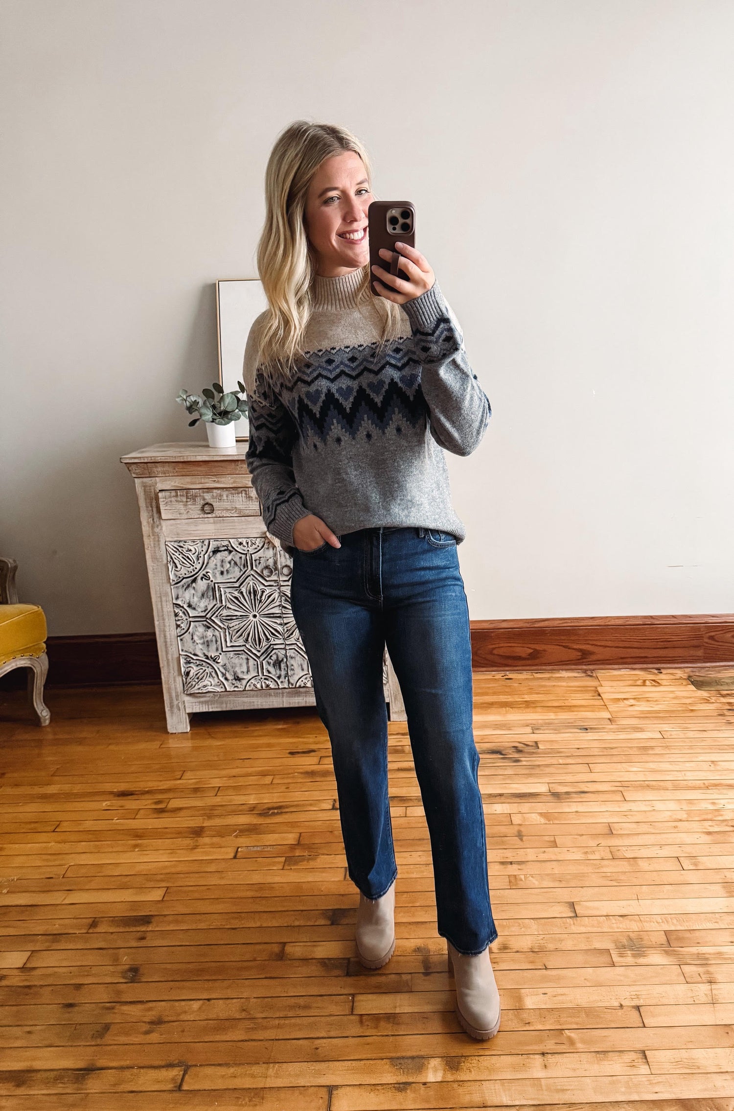 Woman taking a mirror selfie wearing a patterned sweater and blue jeans in a room with wooden flooring.