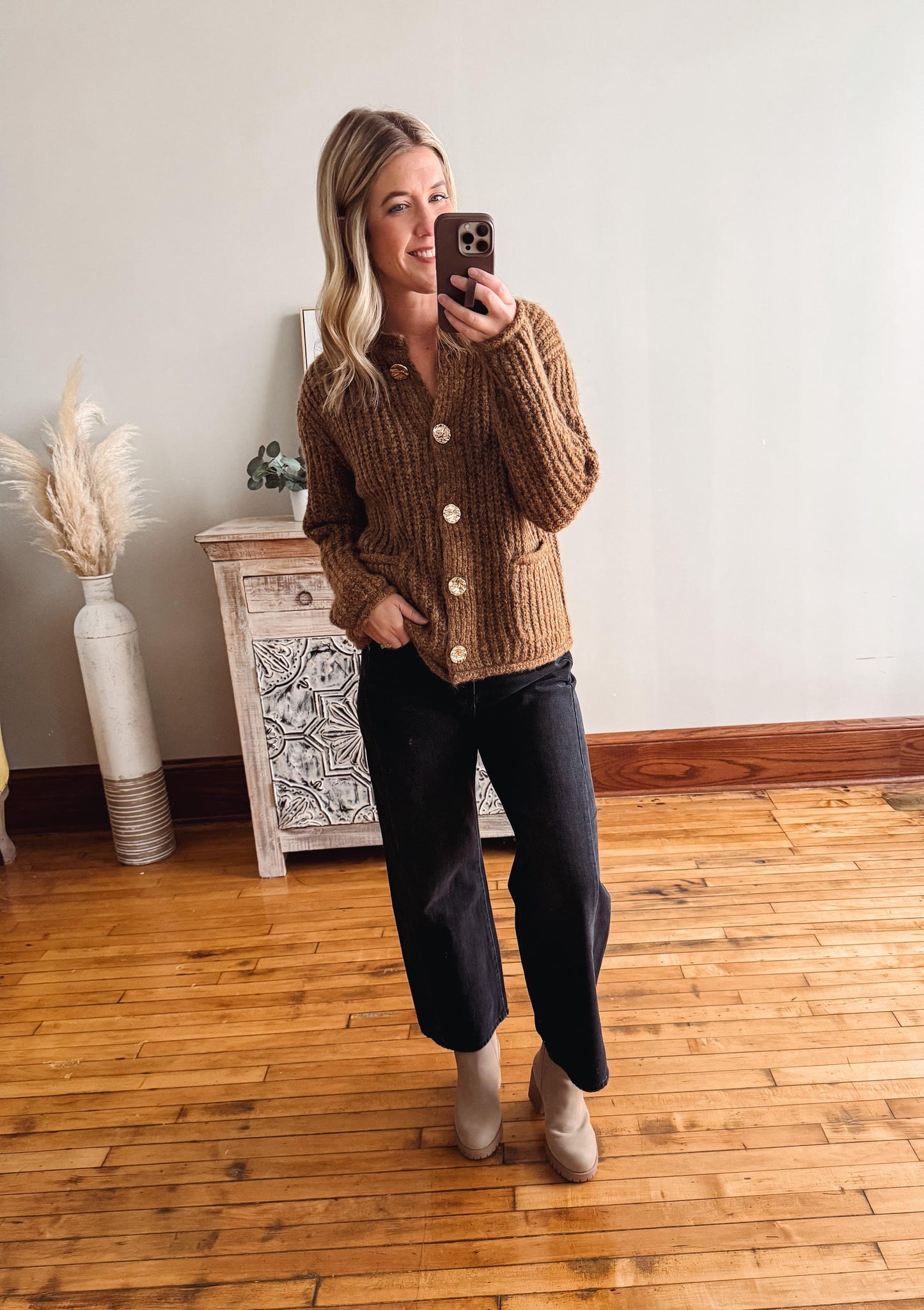 Woman taking a mirror selfie wearing a brown knitted cardigan, black pants, and beige shoes in a room with wooden flooring and a decorative cabinet.