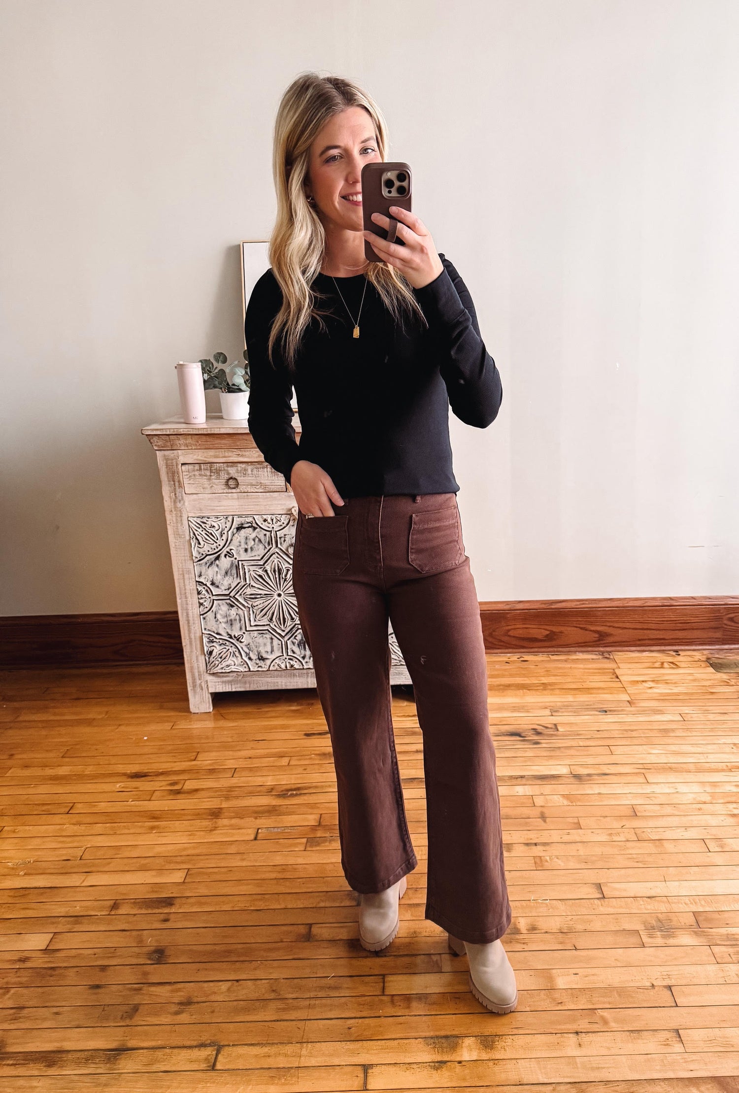 Woman taking a mirror selfie wearing a black top and brown pants in a room with wooden flooring.