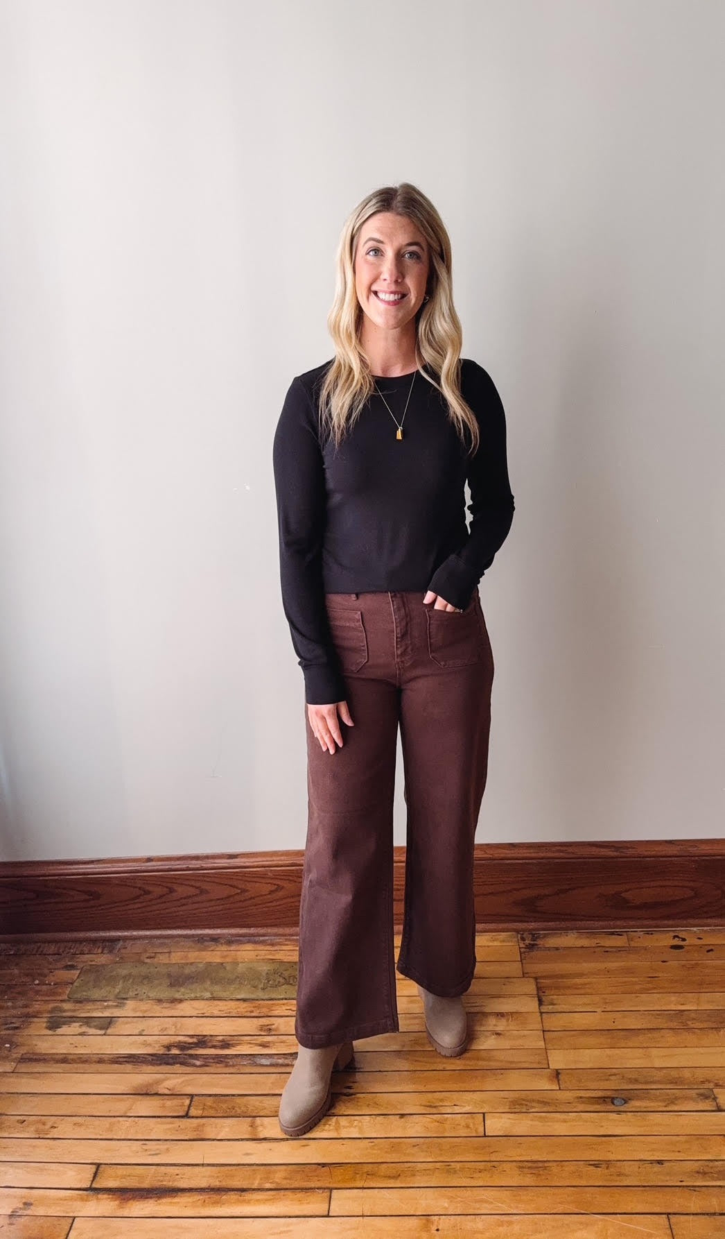 Woman wearing a black top and brown pants standing on a wooden floor with a white wall background