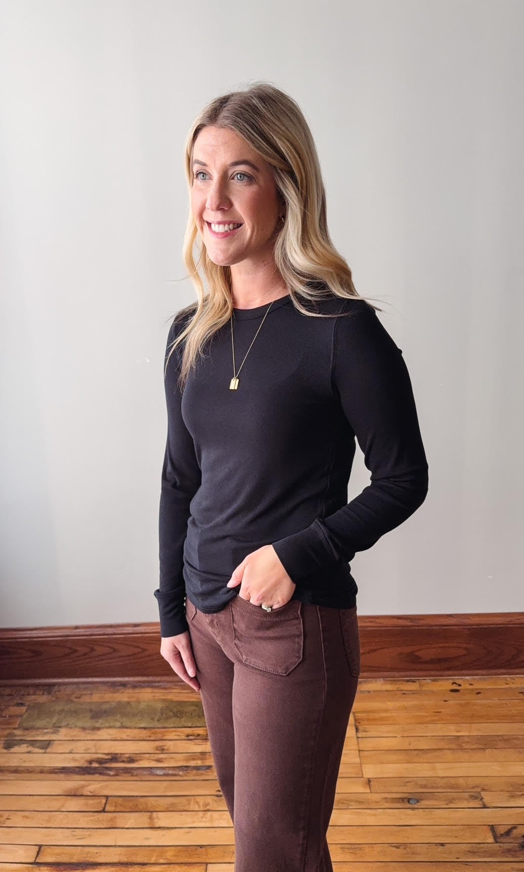 Woman wearing a black long-sleeve top and brown pants standing against a plain wall.