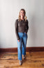 Woman wearing a brown long-sleeve top and blue jeans standing on a wooden floor with a white wall background.