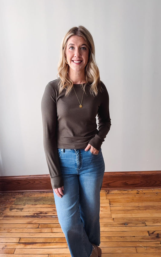 Woman wearing a brown long-sleeve top and blue jeans standing on a wooden floor.