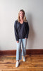 Woman wearing a dark blue sweater and light blue jeans standing on a wooden floor with a white wall background.