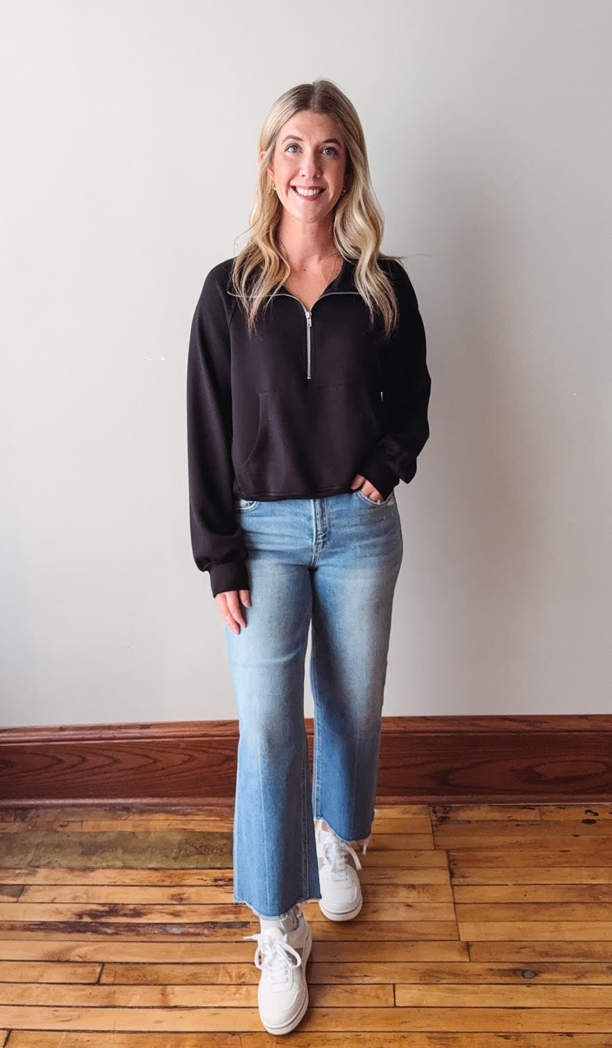 Woman wearing a black sweater and blue jeans standing on a wooden floor.