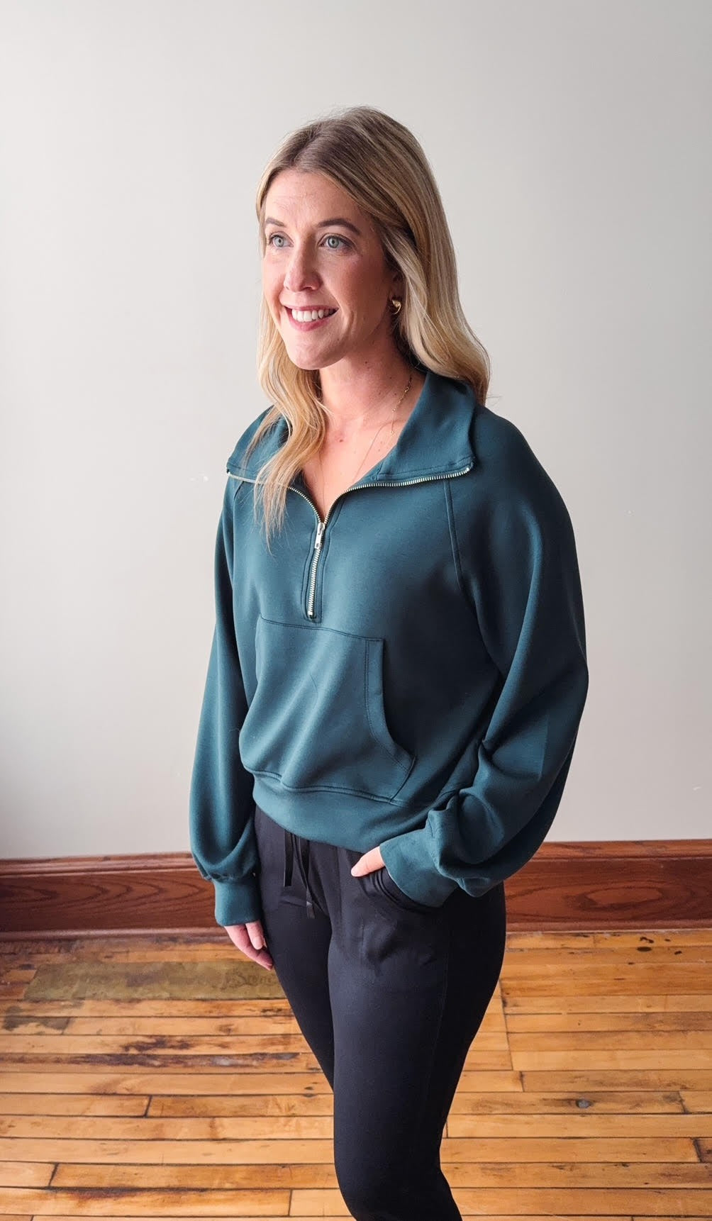 Woman wearing a teal pullover with a zipper on a wooden floor and white wall background