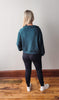 Person wearing a teal sweatshirt and black leggings standing on a wooden floor.