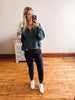 Woman taking a mirror selfie wearing a teal hoodie and black leggings in a room with wooden flooring.