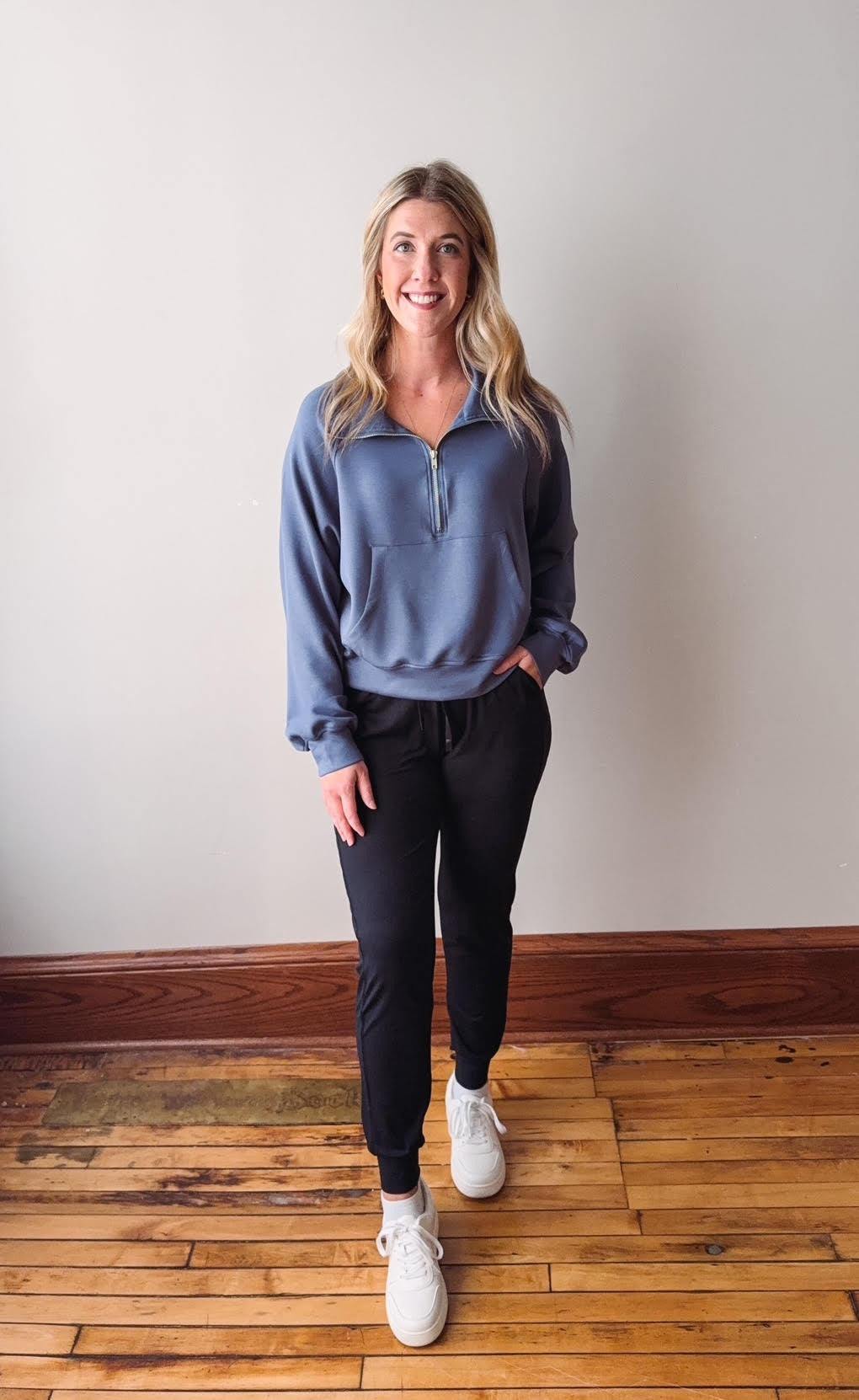 Woman wearing a blue sweater and black pants standing on a wooden floor.