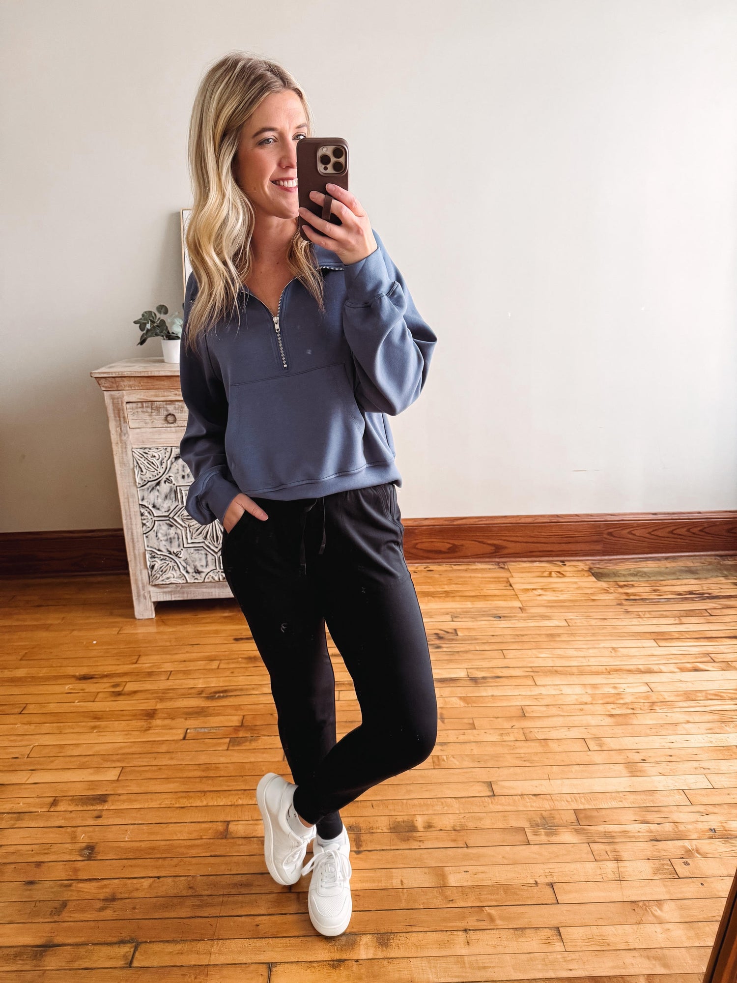 Woman taking a mirror selfie wearing a blue pullover and black pants on a wooden floor.