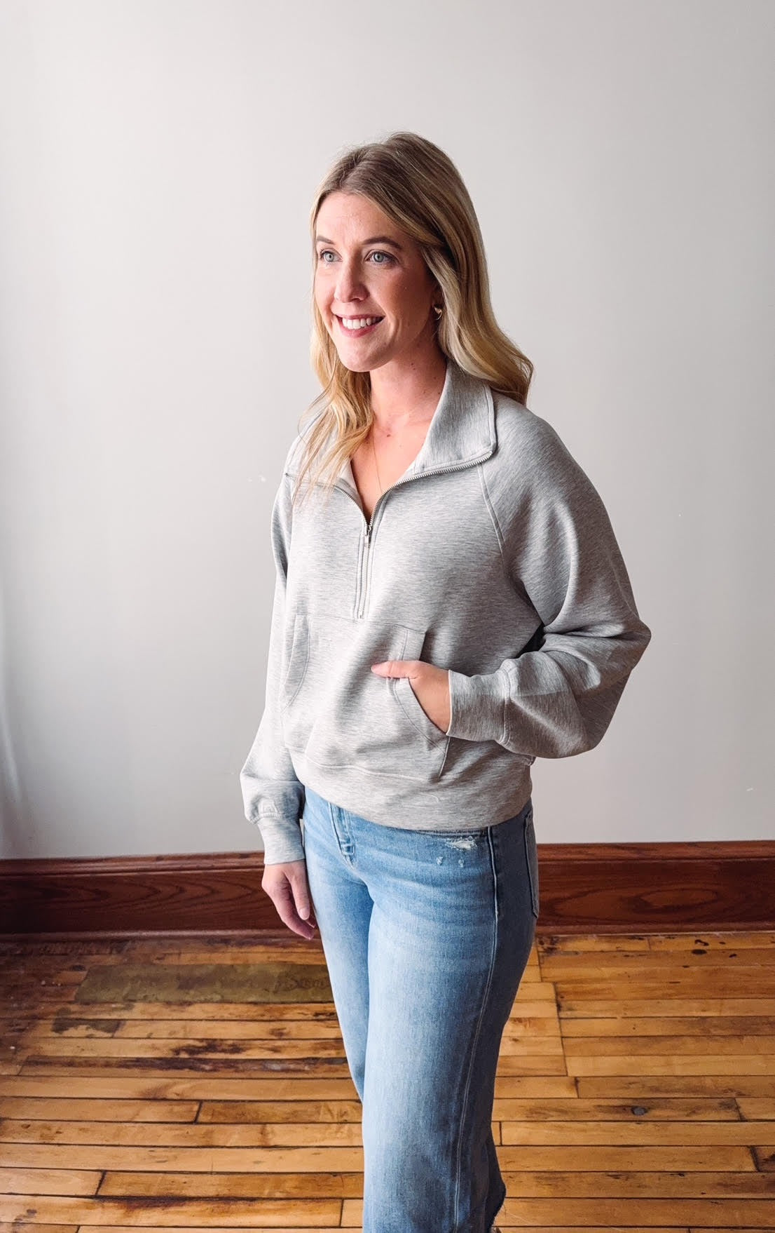 Woman wearing a gray zip-up sweatshirt and light blue jeans standing in a room with wooden flooring and a white wall.