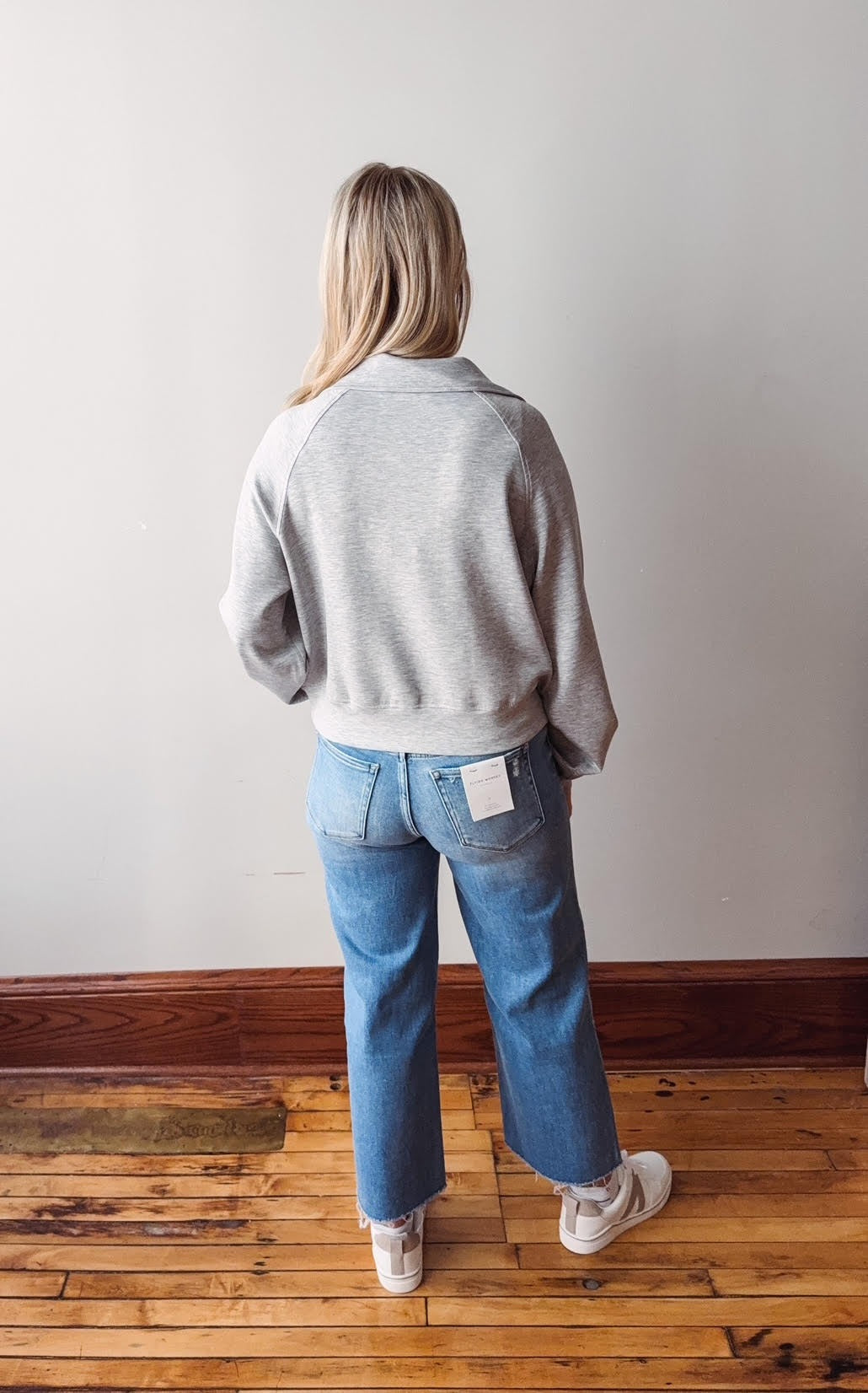 Person wearing a gray sweater and blue jeans standing on a wooden floor with a white wall background