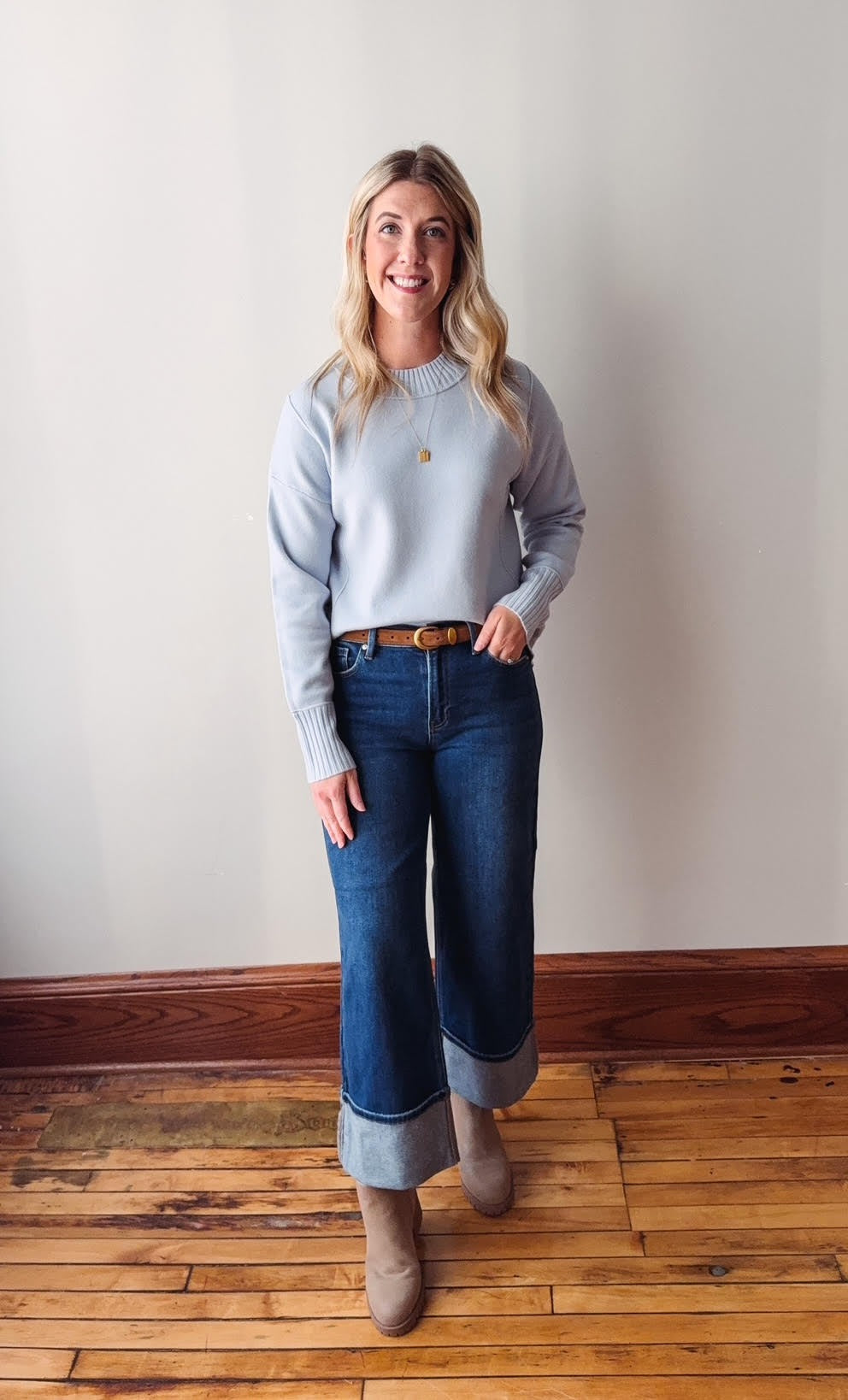 Woman wearing a light blue sweater and blue jeans standing on a wooden floor against a white wall.