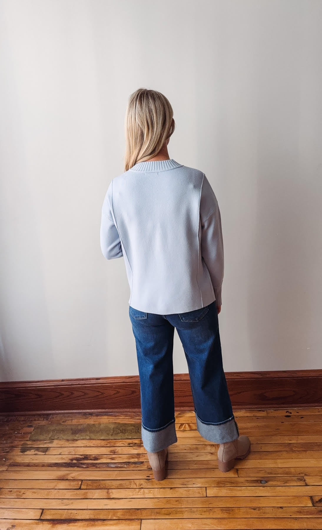 Person wearing a light blue sweater and jeans standing in a room with wooden flooring and a white wall.