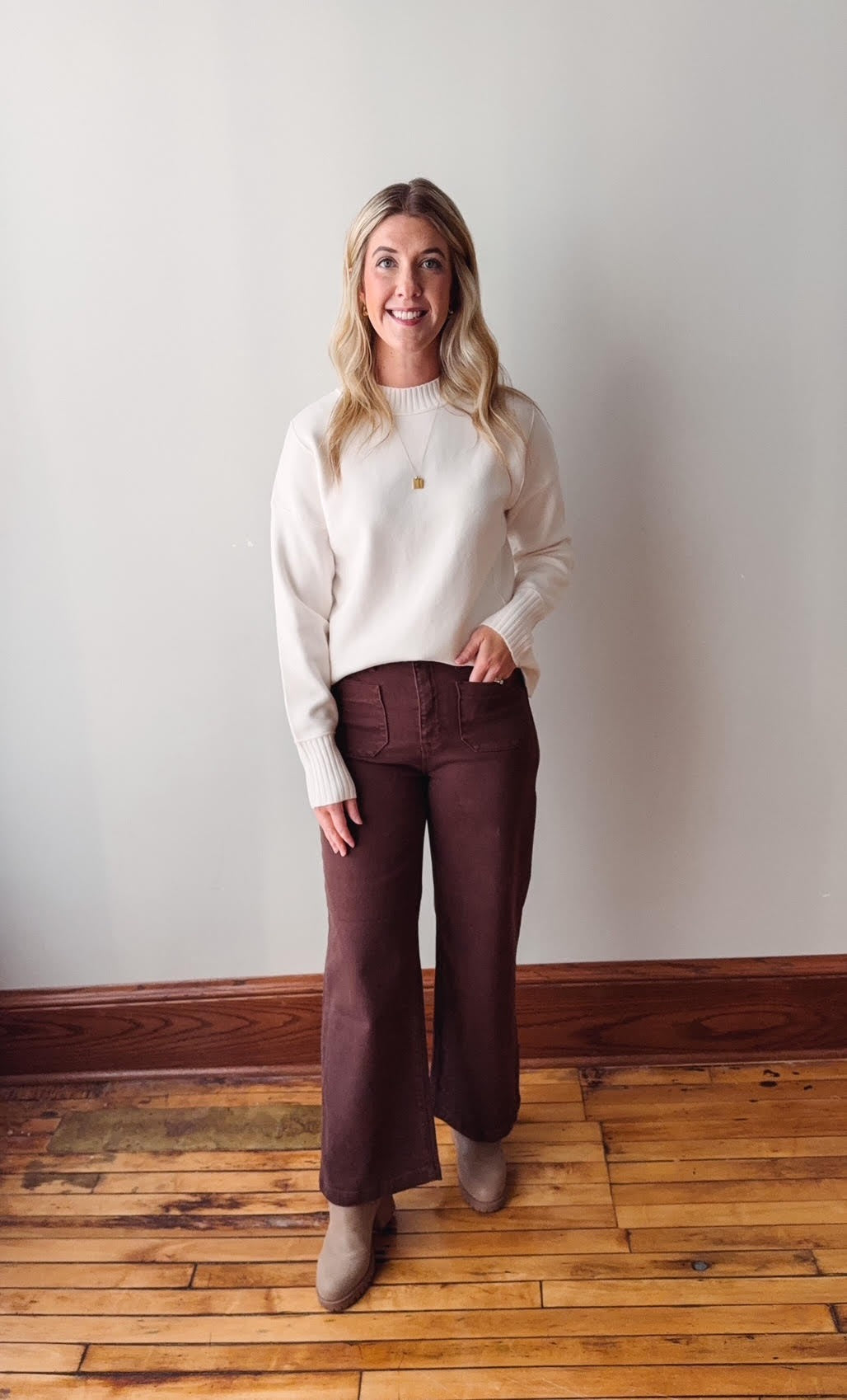 Woman wearing a white sweater and maroon pants standing on a wooden floor against a plain wall.
