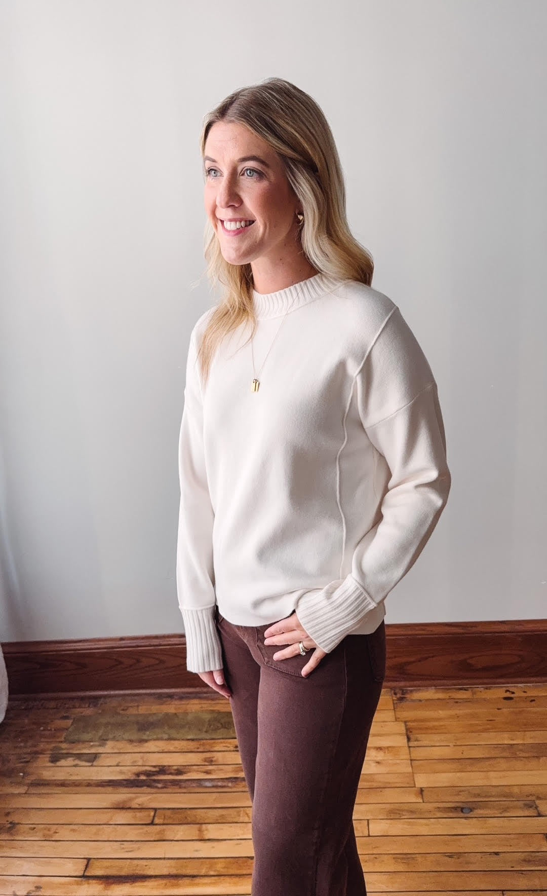 Woman wearing a white sweater and brown pants standing against a plain wall.