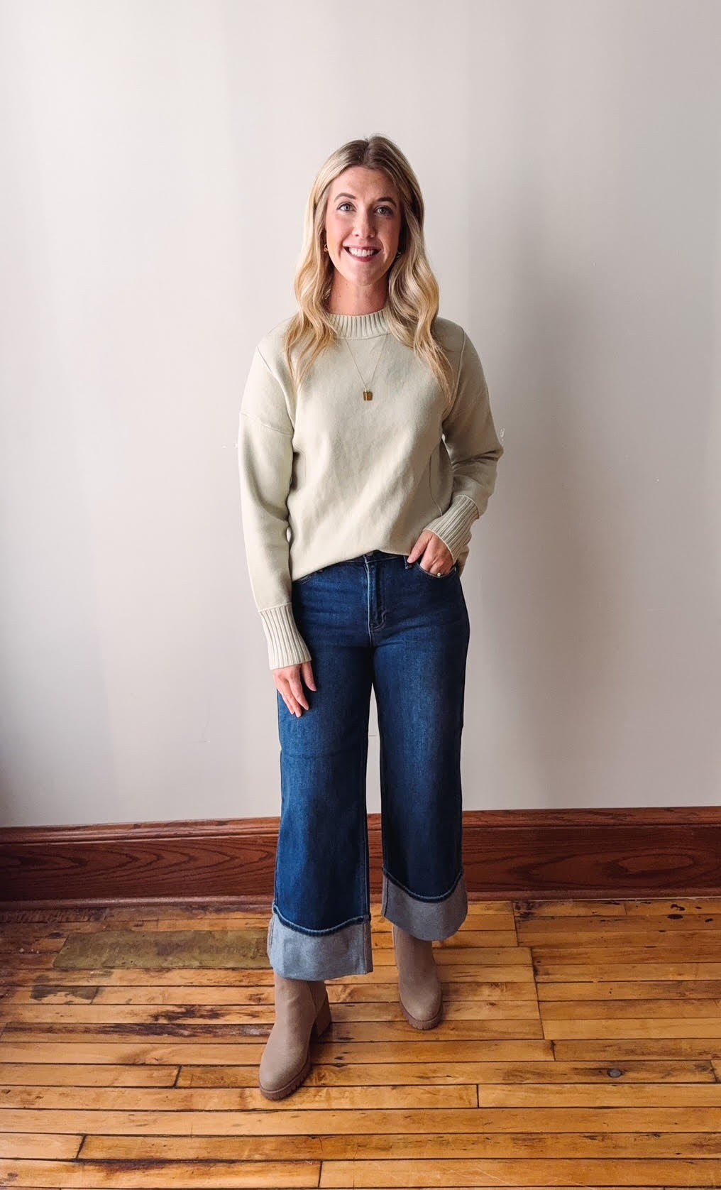 Woman wearing a sage sweater and blue jeans standing on a wooden floor.