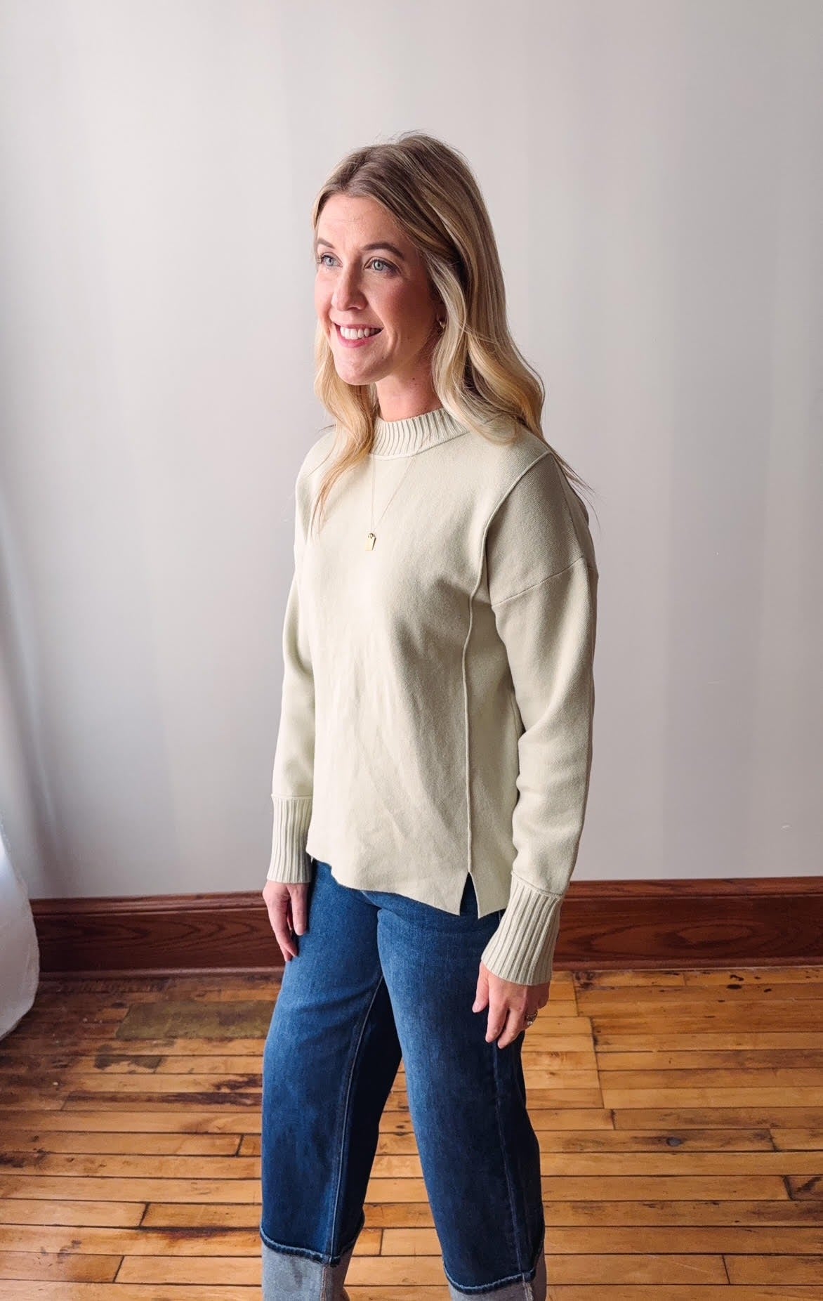 Woman wearing a sage sweater and blue jeans standing in a room with wooden flooring and a white wall.