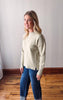 Woman wearing a sage sweater and blue jeans standing in a room with wooden flooring and a white wall.