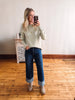 Woman taking a mirror selfie wearing a light green sweater and blue jeans in a room with wooden flooring.