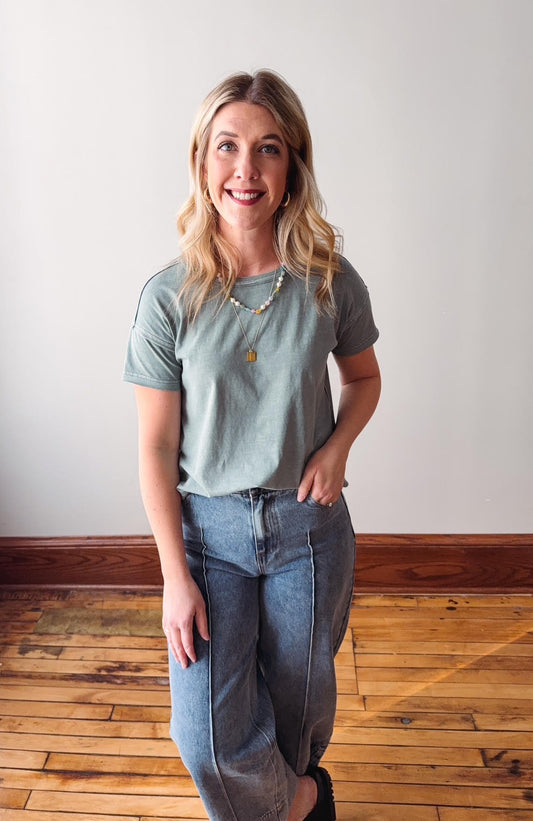 Woman wearing a light blue t-shirt and jeans standing on a wooden floor with a white wall background