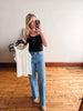 Woman taking a mirror selfie wearing a black top and blue jeans, with a white wall and wooden floor in the background.