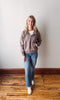Woman wearing a brown sweater and blue jeans standing on a wooden floor with a white wall background