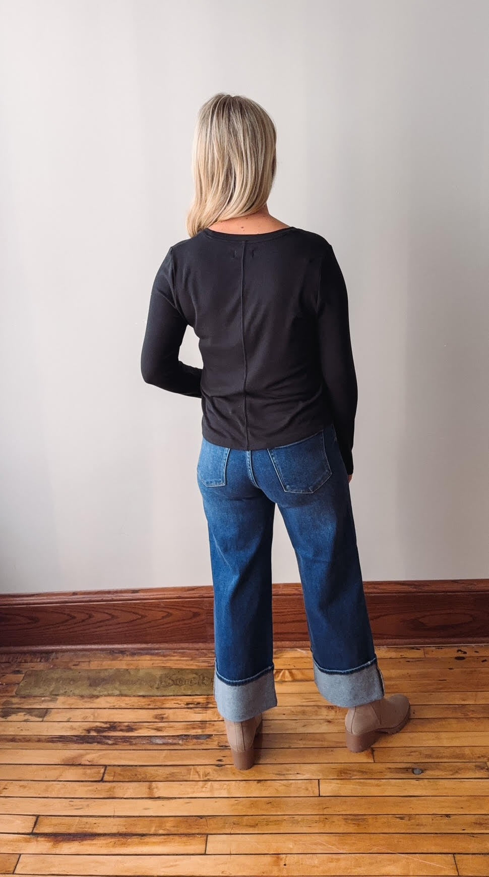 Person wearing a black long-sleeve top and blue jeans standing on a wooden floor with a white wall background.